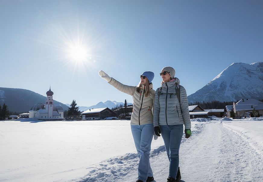 Zwei Frauen wandern im Schnee in der Region Seefeld, mit Kirche und Bergen im Hintergrund.