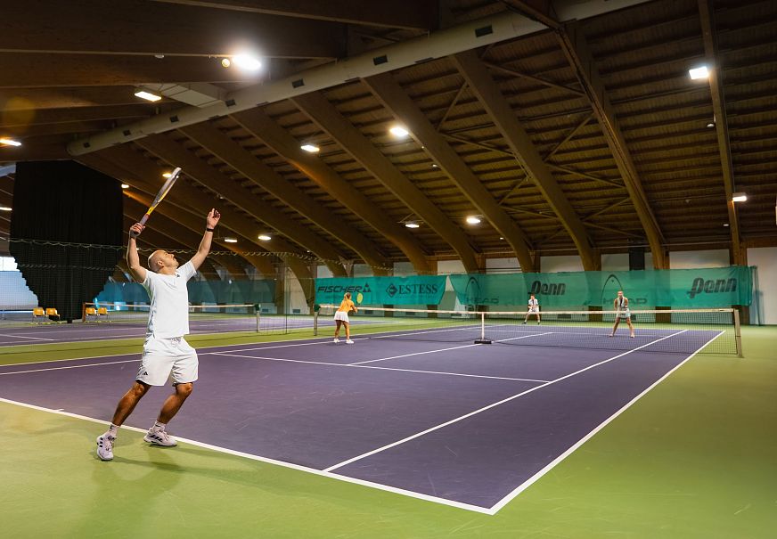 Indoor tennis court with four players; one player serves while others watch.