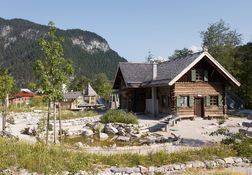 Traditionelle Holzhütte in einer natürlichen Umgebung mit Bergen im Hintergrund und einem liebevoll gestalteten Garten.