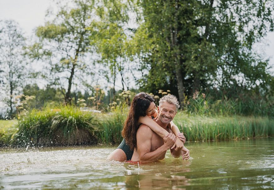 Ein Paar schwimmt in einem Gewässer und umarmt sich, umgeben von grüner Natur.