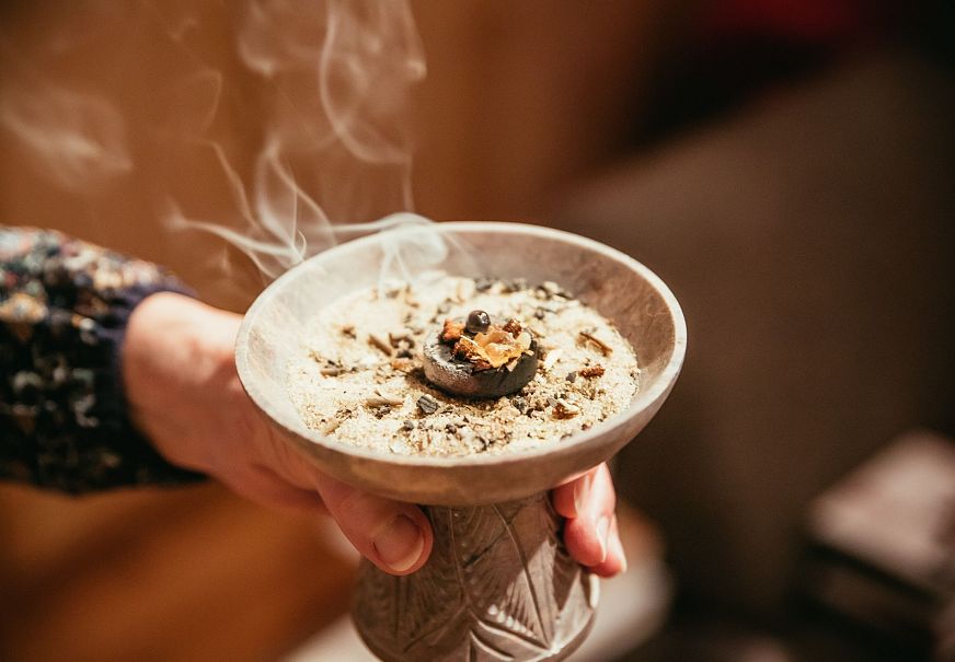 Hand holding a decorative bowl with ash and smoke rising from the material inside.