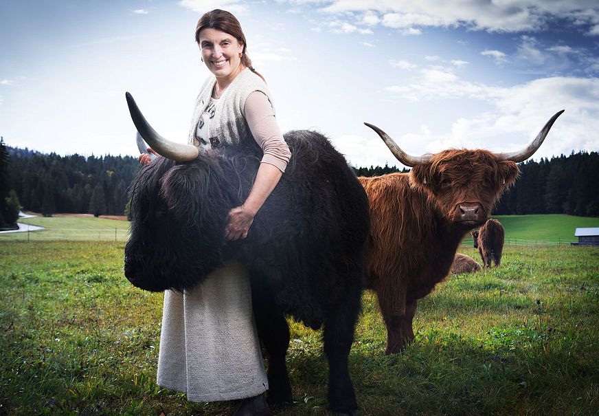 Eine Frau hält ein schwarzes Hochlandrind in einer Wiese, ein braunes Rind steht im Hintergrund.