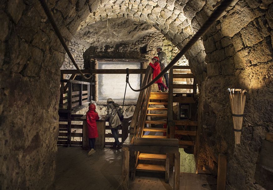 An underground stone chamber with wooden stairs and two figures in red clothing.