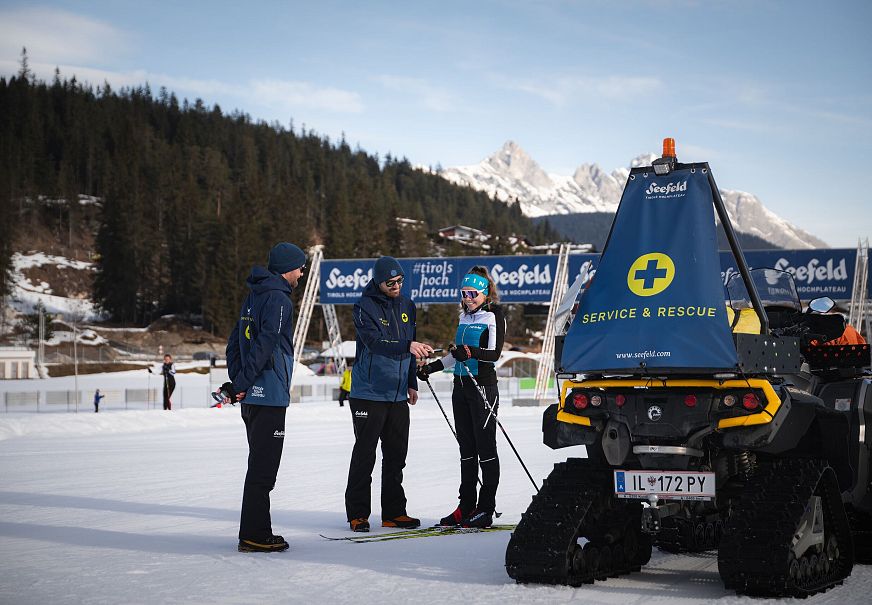 Zwei Männer und eine Skifahrerin besprechen sich vor einem Winterhintergrund mit einem Rettungsfahrzeug.