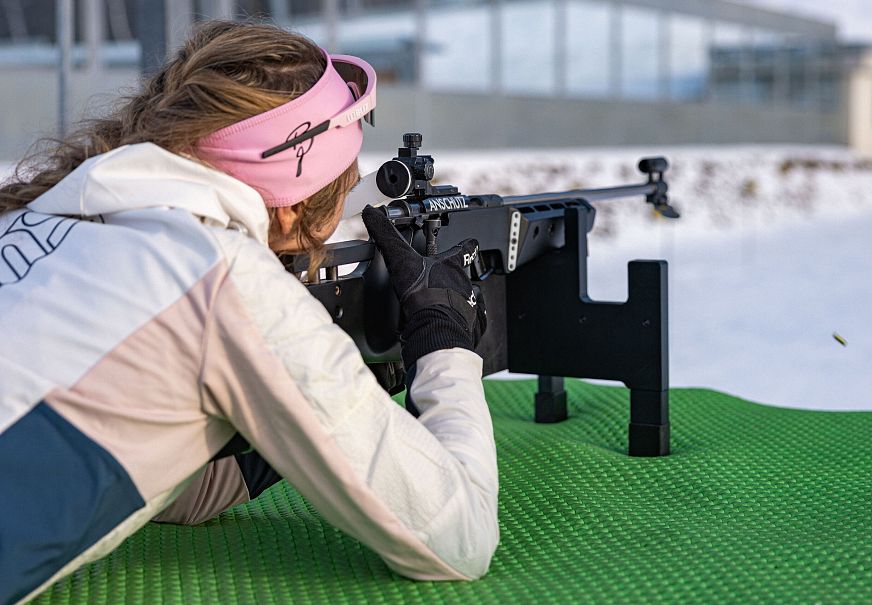 Person zielt mit Biathlongewehr auf Schnee, trägt rosa Stirnband.
