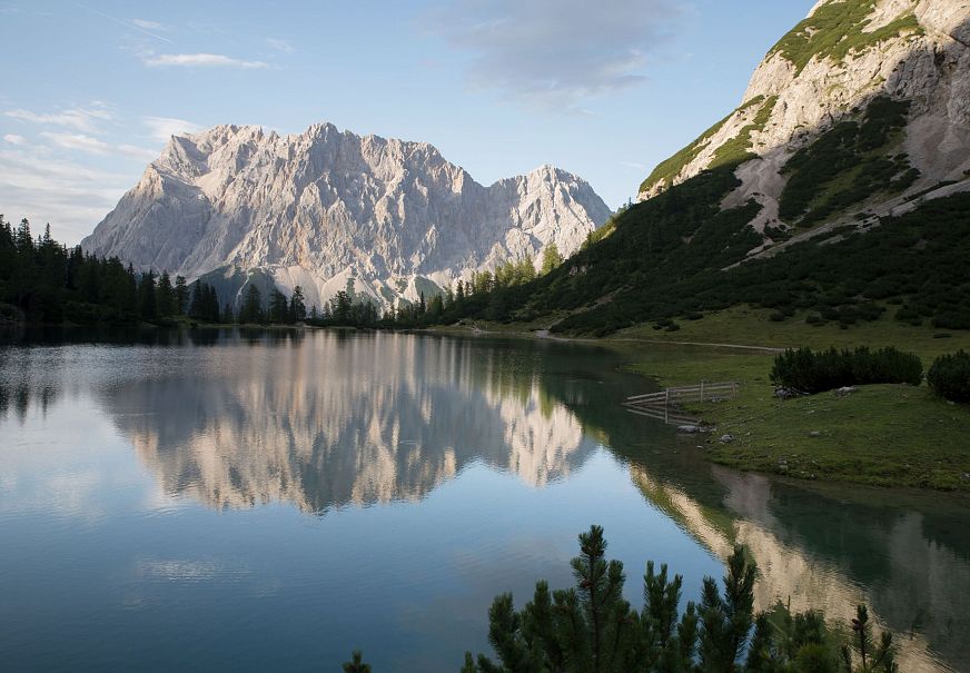 Lakeside view with mountains reflecting in the calm water and greenery nearby.