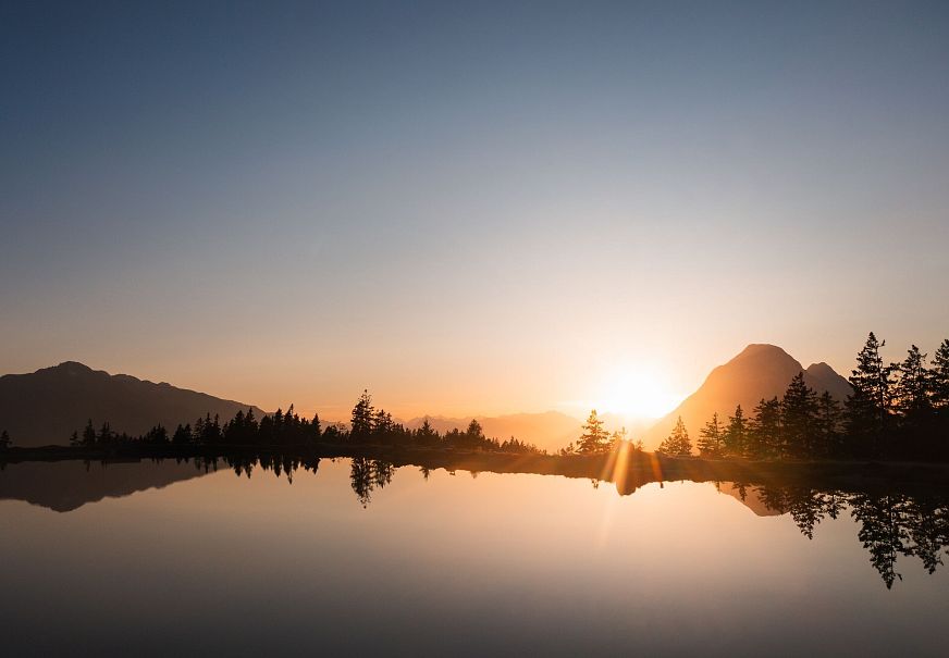 Sonnenaufgang über einem See, umgeben von Bergen und Bäumen.