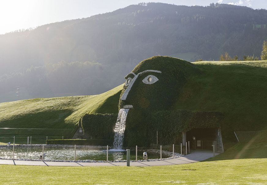 Pflanzenskulptur eines Gesichts mit Wasserfall, umgeben von Hügeln und Bergen.