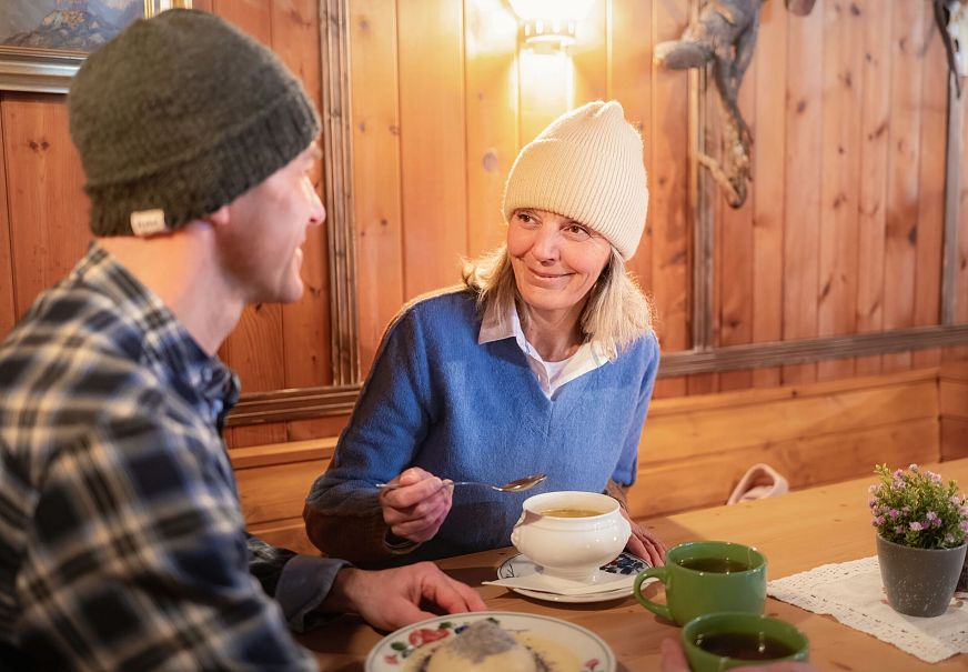 Zwei Personen sitzen in einer Berghütte an einem Tisch mit Essen und Getränken.