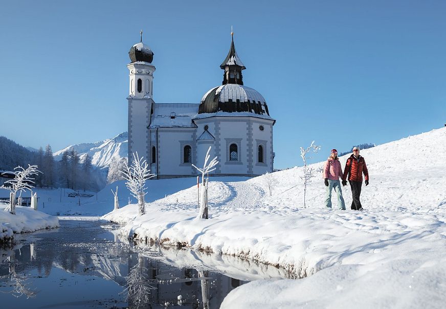 Zwei Wanderer bei einer Winterwanderung vor einer Kirche in einer verschneiten Landschaft.