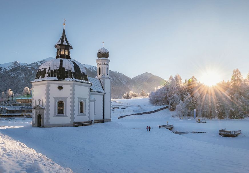 Winterliche Kapelle mit schneebedeckten Bergen und Bäumen im Sonnenlicht in der Region Seefeld.