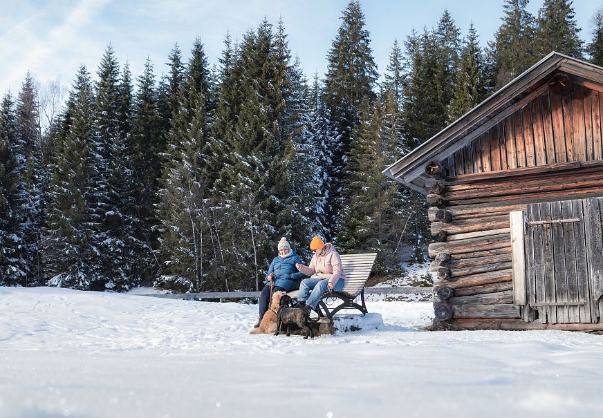Verschneite Landschaft mit Paar auf Bank vor Holzscheune, umgeben von Nadelbäumen.