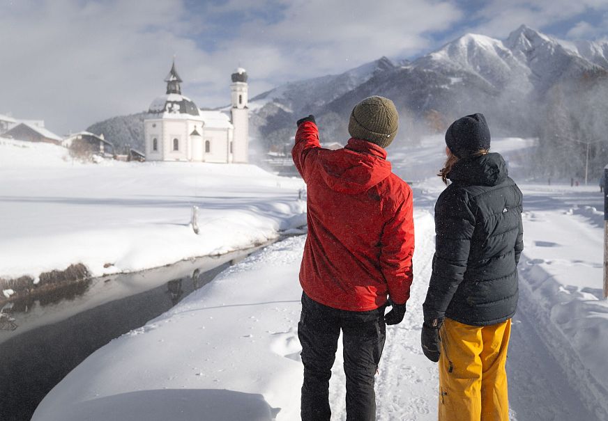 Zwei Personen vor einer verschneiten Kirche in den Alpen, Region Seefeld. Verschneite Landschaft, Berge im Hintergrund.