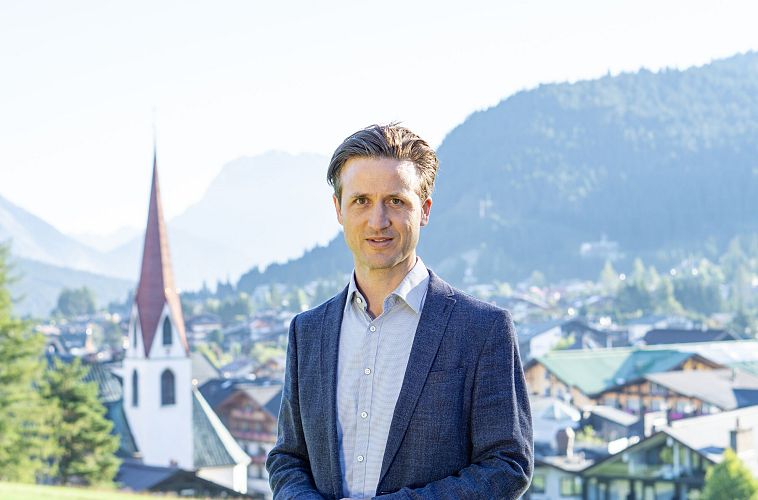 A man in a suit stands with mountains and a village in the background.