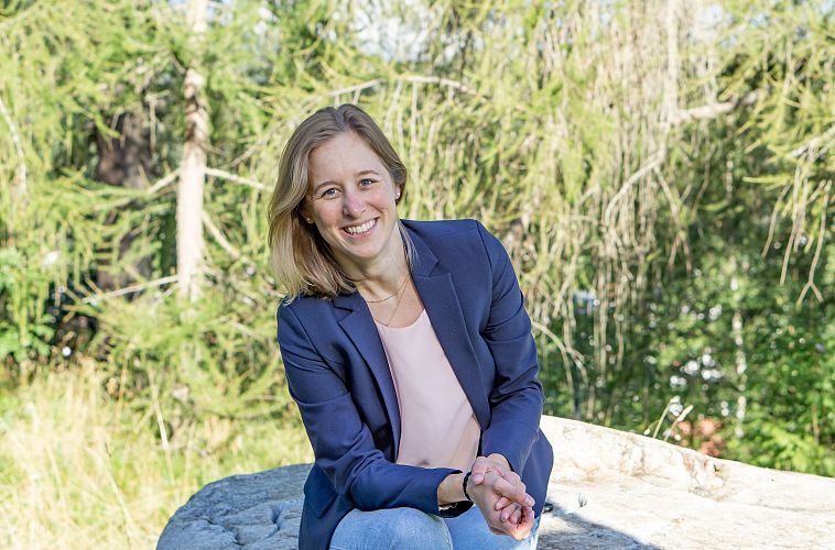 Eine Frau mit Blazer sitzt lächelnd auf einem Felsen in der Natur.