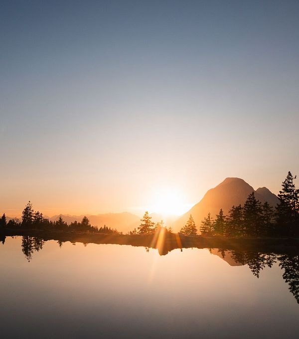 Sonnenaufgang über einem See, umgeben von Bergen und Bäumen.