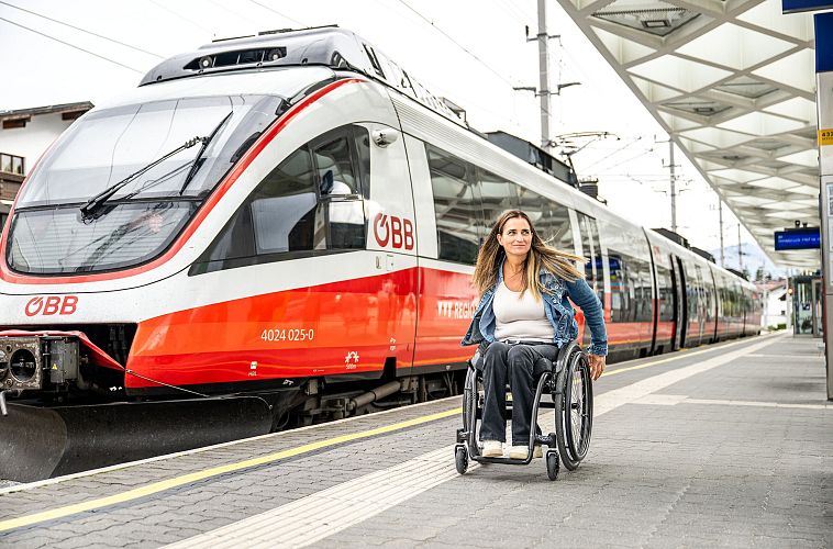 Frau im Rollstuhl an Bahnsteig mit einfahrendem ÖBB-Zug im Hintergrund.