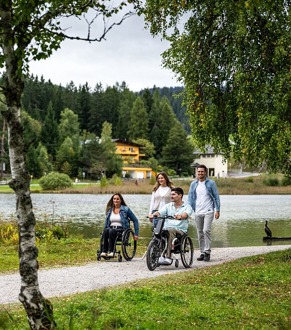 Familie am Seeweg in der Region Seefeld, zwei Personen in Rollstühlen, malerische Umgebung mit Bäumen und bewölktem Himmel.