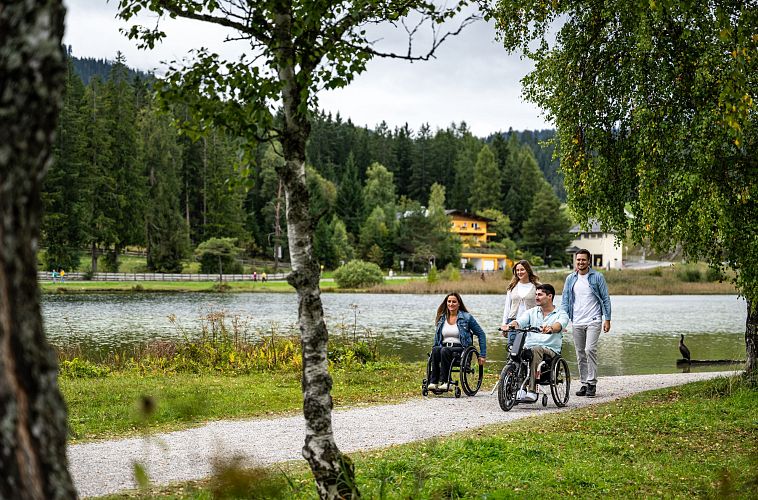Familie am Seeweg in der Region Seefeld, zwei Personen in Rollstühlen, malerische Umgebung mit Bäumen und bewölktem Himmel.