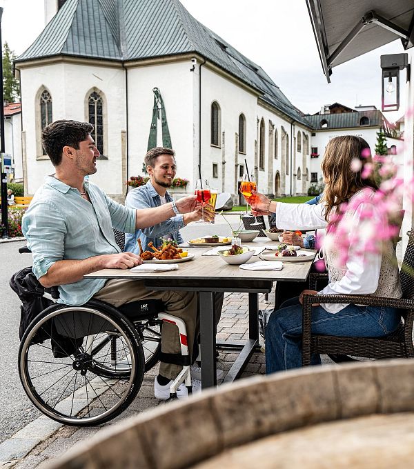 Drei Personen essen draußen in einem Bergdorf Nähe der Region Seefeld, mit einer Kirche im Hintergrund. Eine Person sitzt im Rollstuhl.