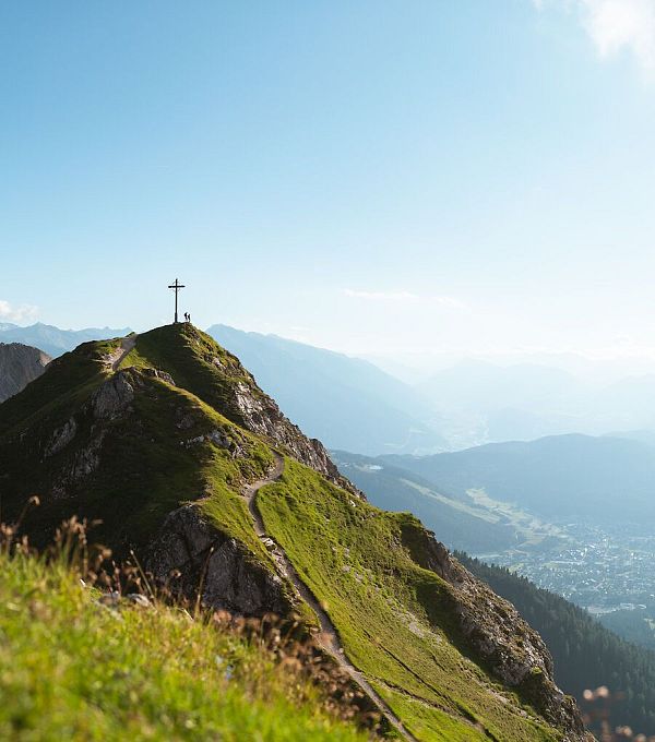 Berglandschaft mit einem Kreuz auf einem Hügel und schroffen Bergen im Hintergrund.