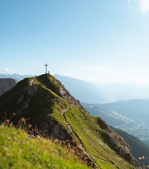 Berglandschaft mit einem Kreuz auf einem Hügel und schroffen Bergen im Hintergrund.