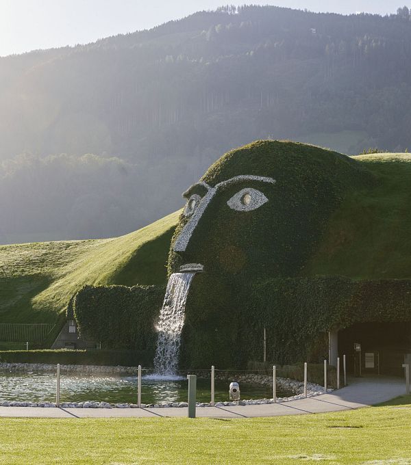 A large grassy face sculpture with waterfall, surrounded by hills in soft sunlight.