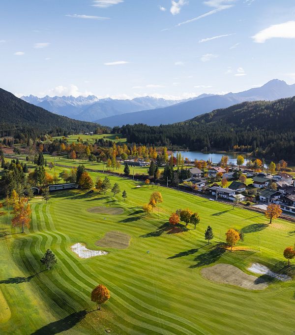 Landschaft mit Golfplatz, Bergen, buntem Laub und kleinen Häusern im Hintergrund.
