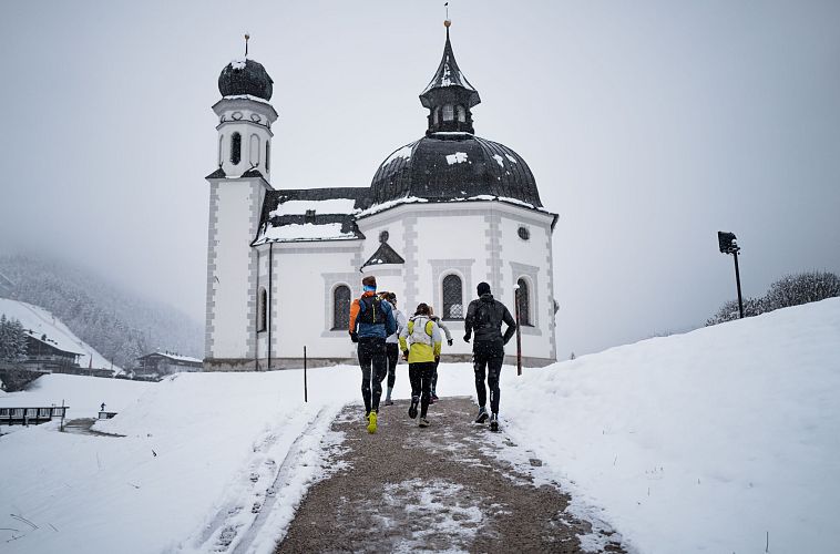Läufergruppe vor verschneiter Kirche mit Zwiebelturm, Winterlauf in der Region Seefeld.