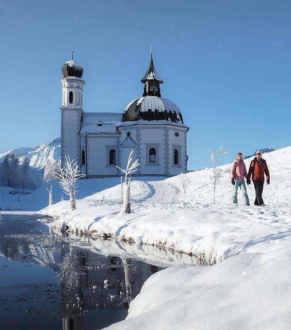 Verschneite Kirche in Winterlandschaft, Wanderer im Vordergrund. Region Seefeld. Klarer Himmel, blau.