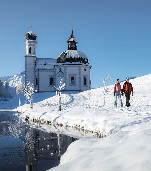 Zwei Wanderer bei einer Winterwanderung vor einer Kirche in einer verschneiten Landschaft.
