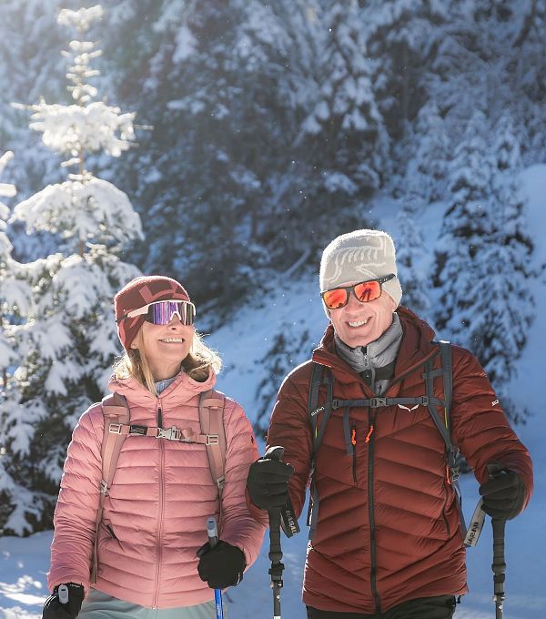 Paar beim Winterwandern im Schnee, mit Jacken und Mützen, umgeben von schneebedeckten Bäumen, in sonniger Winterlandschaft.