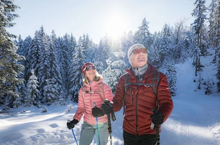 Winterwandern in der Region Seefeld_Wildmoos_Mann und Frau laufen durch den Wald im Wildmoos und blicken in die Bäume_Nahaufnahme von vorne (1)