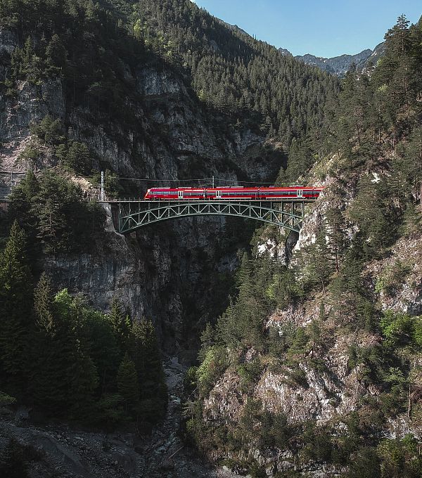 Red train on a green bridge surrounded by mountains and trees.