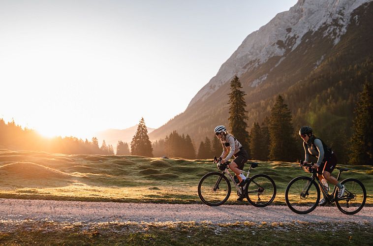 Zwei Radfahrer fahren im Sonnenaufgang durch eine bewaldete Landschaft.