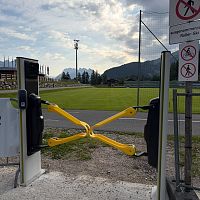 Zugang mit Drehkreuz an einem Fußballplatz, im Hintergrund Berge und grüner Rasen.