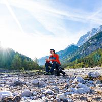 Eine Person sitzt auf einem Kiesfeld vor einer Berglandschaft mit strahlender Sonne.