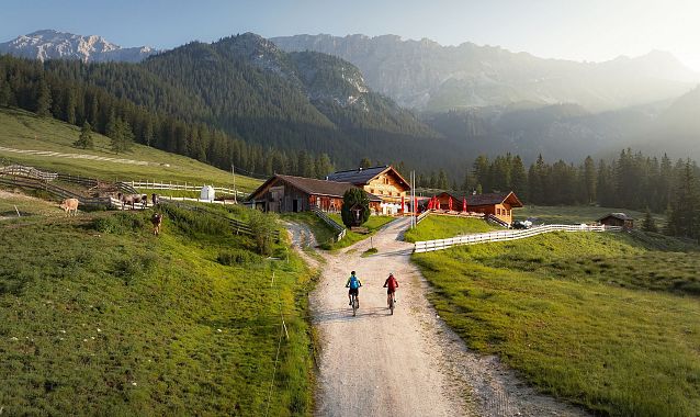 Two cyclists riding along a gravel path towards a lodge in a mountainous landscape.