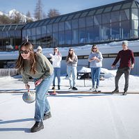 Gruppe spielt Eisstockschießen in der Region Seefeld vor modernem Gebäude auf schneebedeckter Eisfläche.