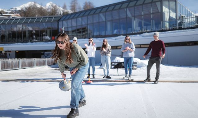 Gruppe spielt Eisstockschießen in der Region Seefeld vor modernem Gebäude auf schneebedeckter Eisfläche.