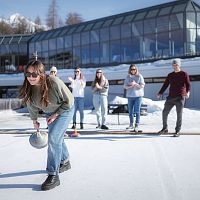 Gruppe spielt Eisstockschießen in der Region Seefeld vor modernem Gebäude auf schneebedeckter Eisfläche.