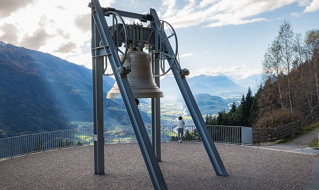 A bell on a metal frame with a scenic mountain view and a person standing nearby.