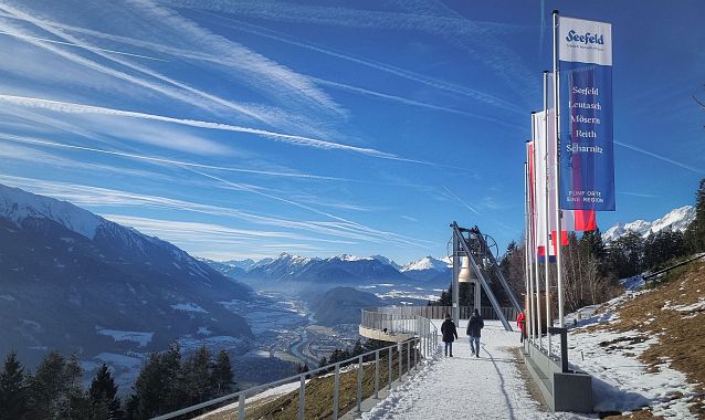 Spaziergänger auf verschneitem Weg bei der Friedensglocke Mösern, Region Seefeld, umgeben von Alpen und klarem Himmel.