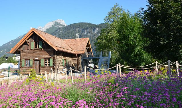 Ein Holzhaus umgeben von Blumen und Bäumen mit Bergen im Hintergrund.