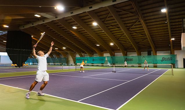 Tennishalle, Spieler in weiß serviert. Weitere Spieler im Hintergrund aktiv. Helle Beleuchtung. Innenaufnahme.