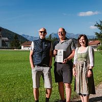 Drei Personen auf einer Wiese vor einem Kirchengebäude und Bergen im Hintergrund.