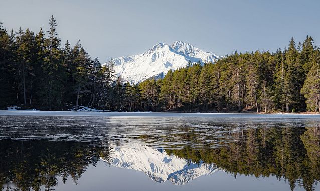 Berglandschaft mit schneebedecktem Gipfel und Wald, der sich in einem ruhigen See spiegelt.