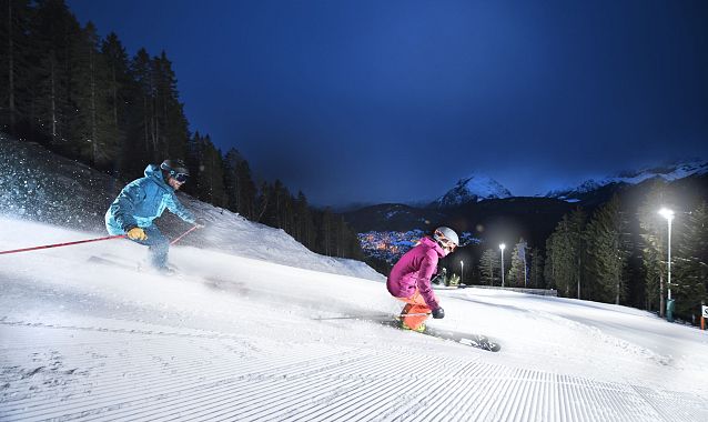 Zwei Skifahrer bei Nacht auf beleuchteter Piste durch verschneiten Wald. Nacht-Skifahren in der Region Seefeld.
