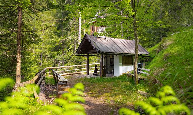 Holzkapelle im Wald mit umgebenden Bäumen und einer Bank.