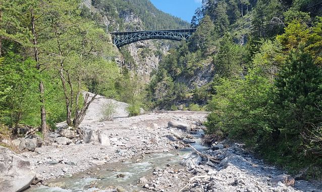 View of a rocky riverbed with a stream and a bridge above in a mountainous area.